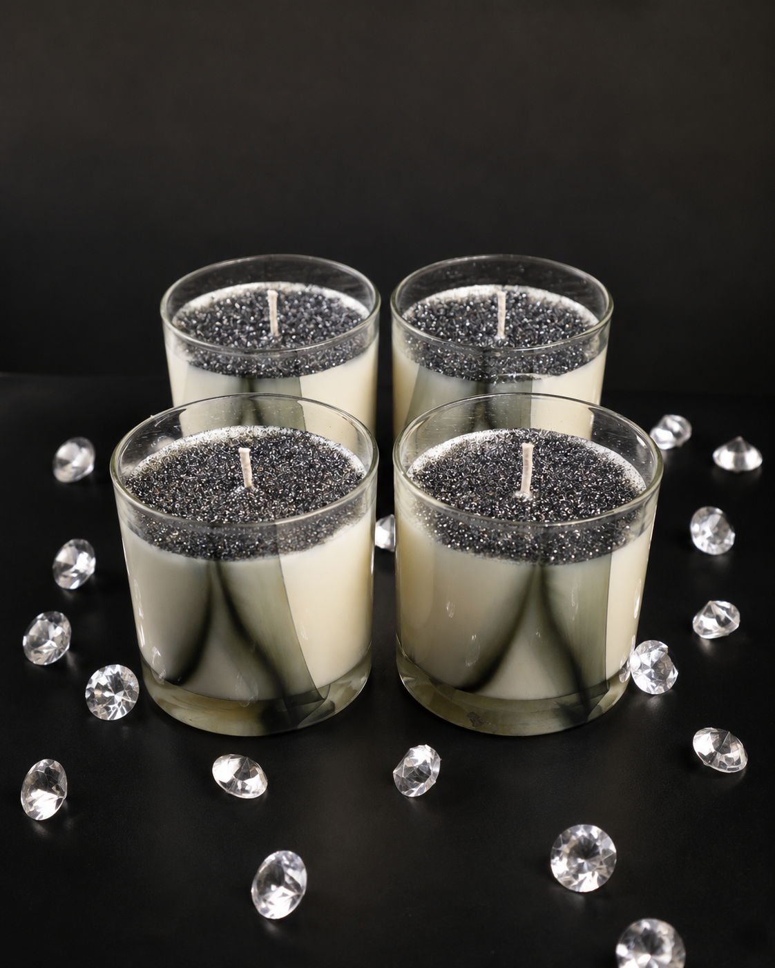 Four glass candles with glittery bases on a black surface with decorative crystals.