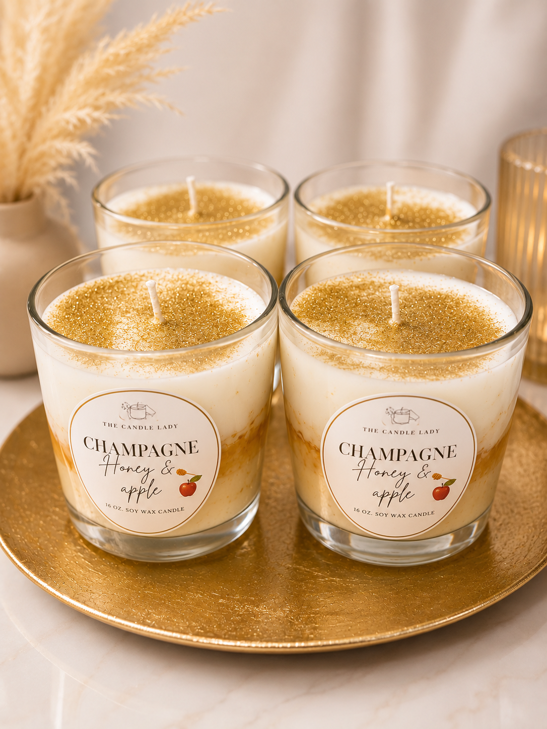 Four glass jar candles with white wax and gold glitter on top, arranged on a round gold tray. The front two jars have round labels reading 'CHAMPAGNE Honey & apple' and 'THE CANDLE LADY'.