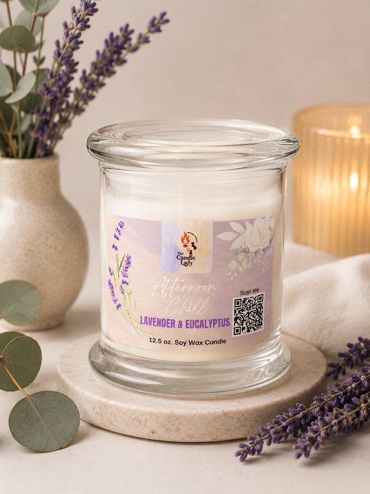 A glass jar candle labeled 'Afternoon Chill' and 'Lavender & Eucalyptus' sits on a round stone coaster, styled with lavender sprigs, a ceramic vase with greenery, and a softly lit candle in the background.