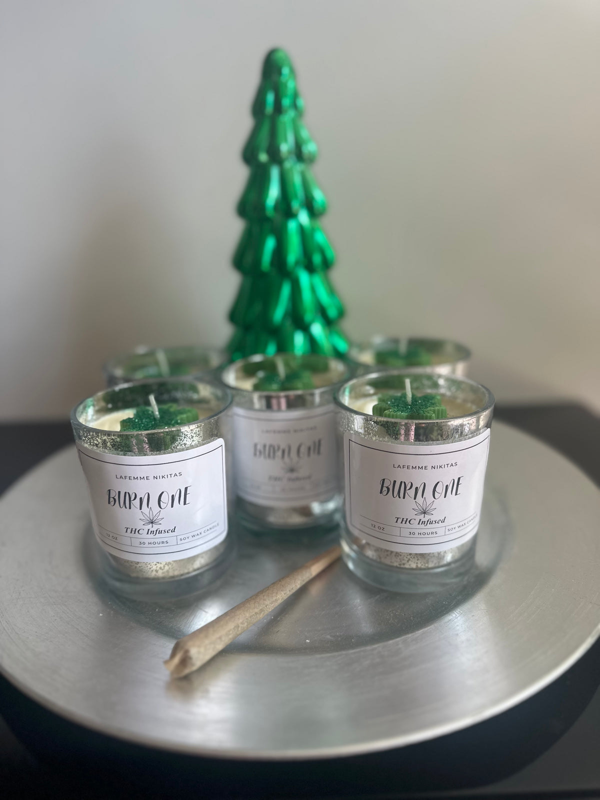 Three silver jar candles labeled Burn One, alongside a green decorative tree, and hand rolled blunt all presented on a silver tray.