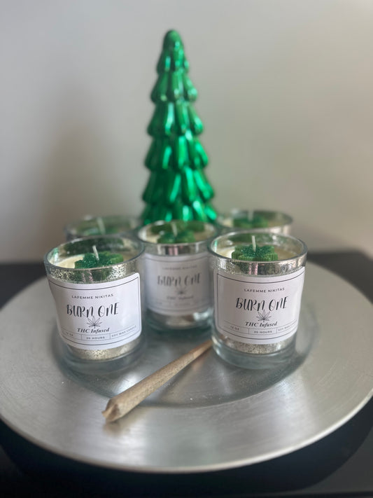 Three silver jar candles labeled Burn One, alongside a green decorative tree, and hand rolled blunt all presented on a silver tray.