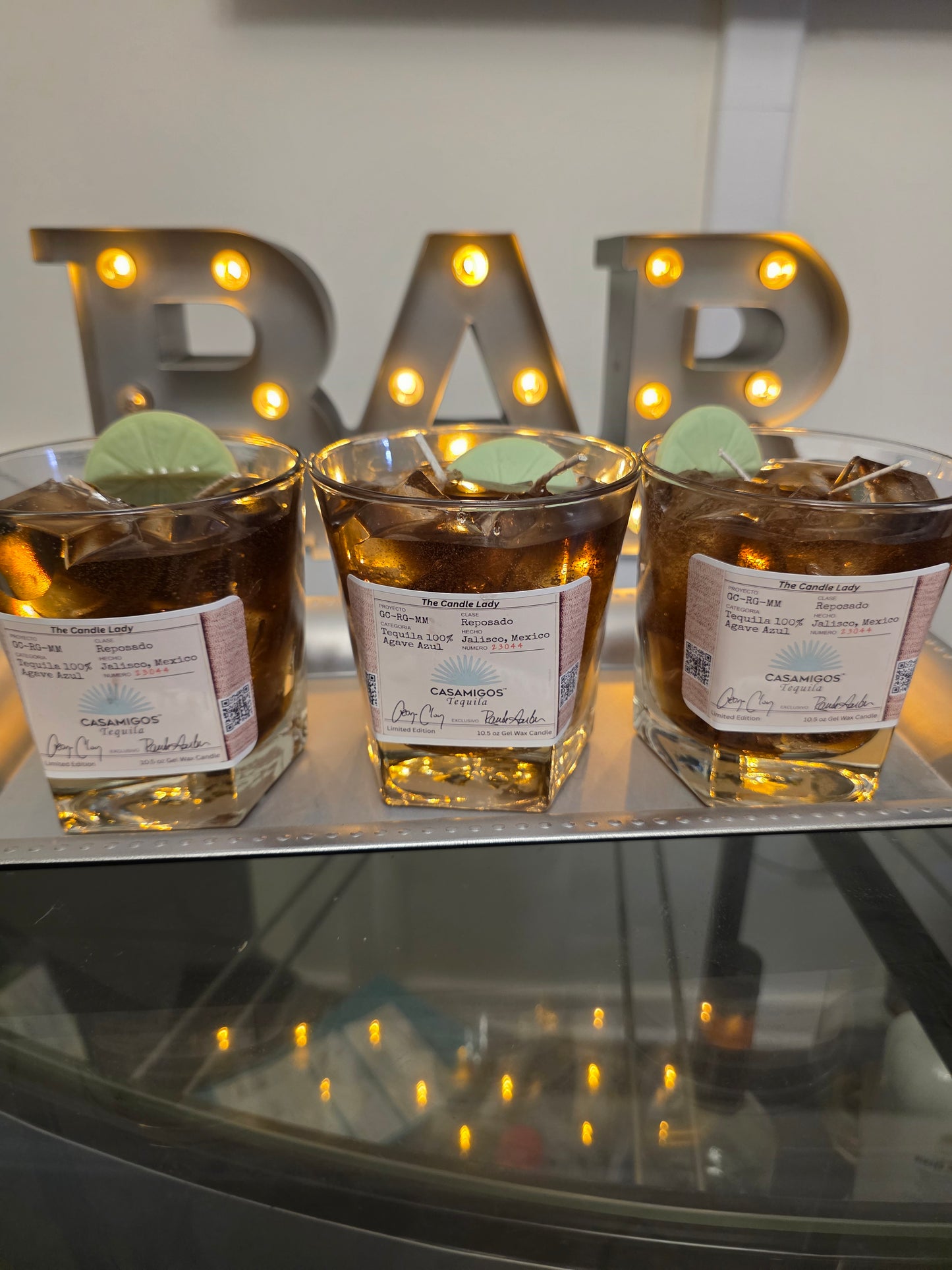 Three casamigos candles with labels on a reflective surface with decorative lights in the background