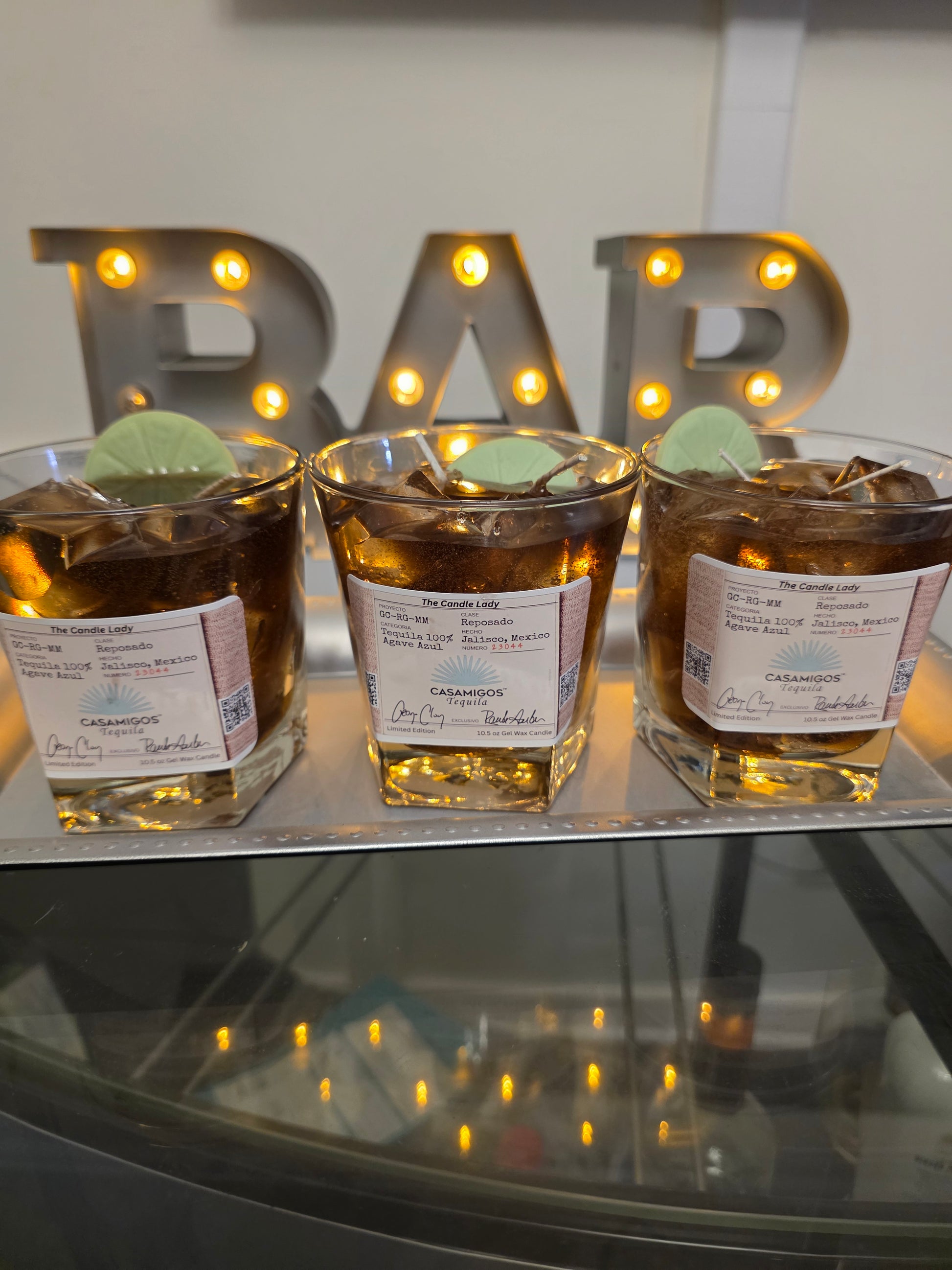 Three casamigos candles with labels on a reflective surface with decorative lights in the background