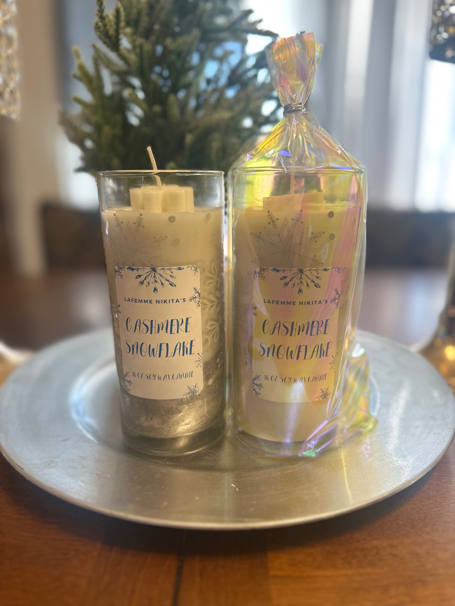 Two 16 oz soy wax candles in Tall snowflake glasses labeled Cashmere Snowflake sitting on a silver tray in front of small Christmas tree