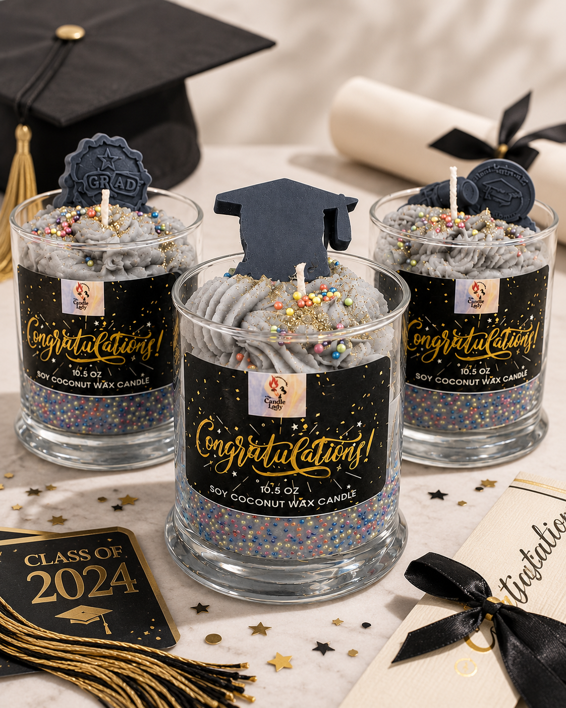 Three graduation-themed soy coconut wax candles in clear glass jars with black labels and gold script text, topped with gray whipped wax, sprinkles, and dark blue graduation cap toppers. A black graduation cap, diploma, and confetti are arranged around them on a light surface.
