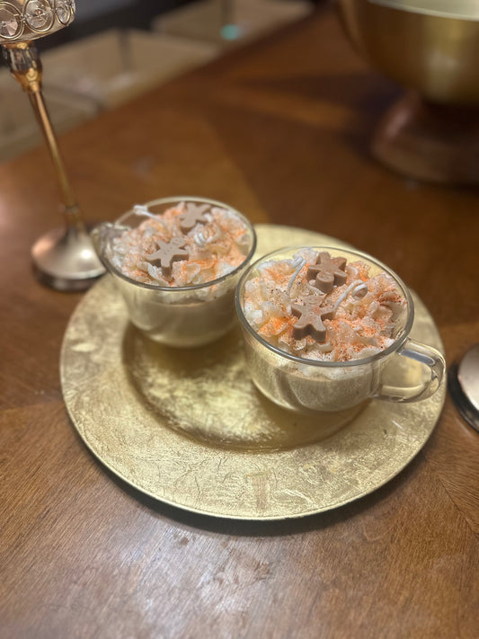 Two clear glass mugs made with soy wax topped with gingerbread-shaped men on a gold coaster on a wooden surface.
