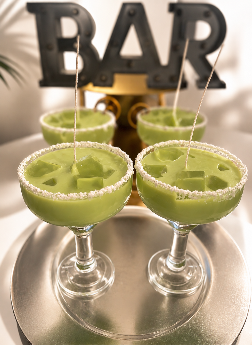 Three green cocktail inspired soy wax candles in margarita-style glasses with sugar-rimmed edges, arranged on a round silver tray with a decorative black and gold BAR sign in the background.