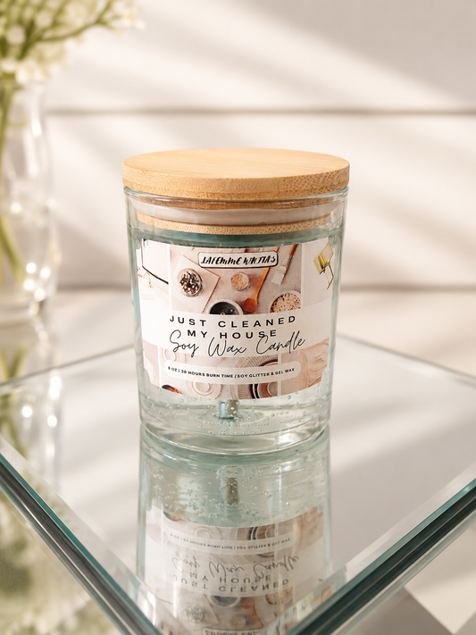 A glass jar candle with a bamboo wood lid sits on a glass shelf. The candle features a white label with the scent name 'Just Cleaned My House' and illustrations of cleaning supplies.