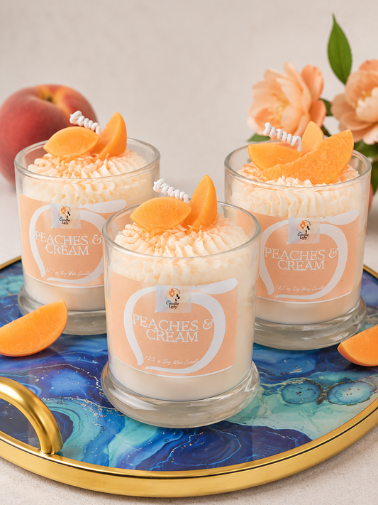 Three candles in clear glass jars labeled 'Peaches & Cream' are arranged on a blue and gold tray with peach slices and flowers. The candles have pale peach and cream wax with whipped cream-like tops and decorative peach wedges.