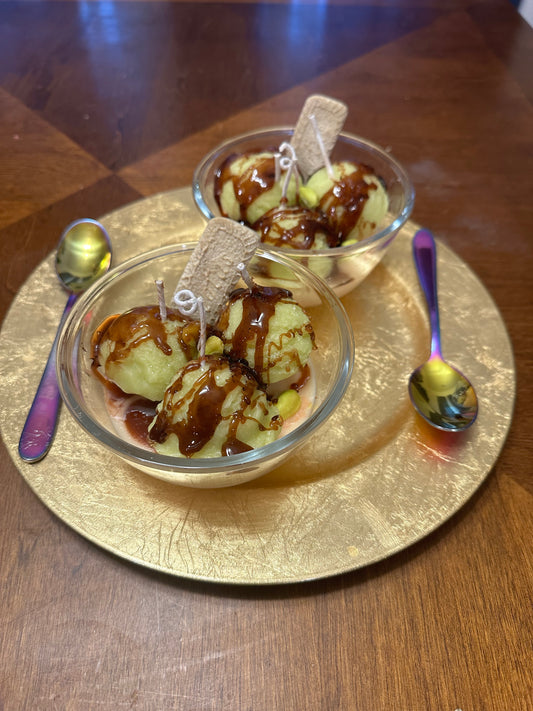 Two glass bowls of ice cream made with soy wax, topped with chocolate drizzle and cookies on a gold plate with spoons.