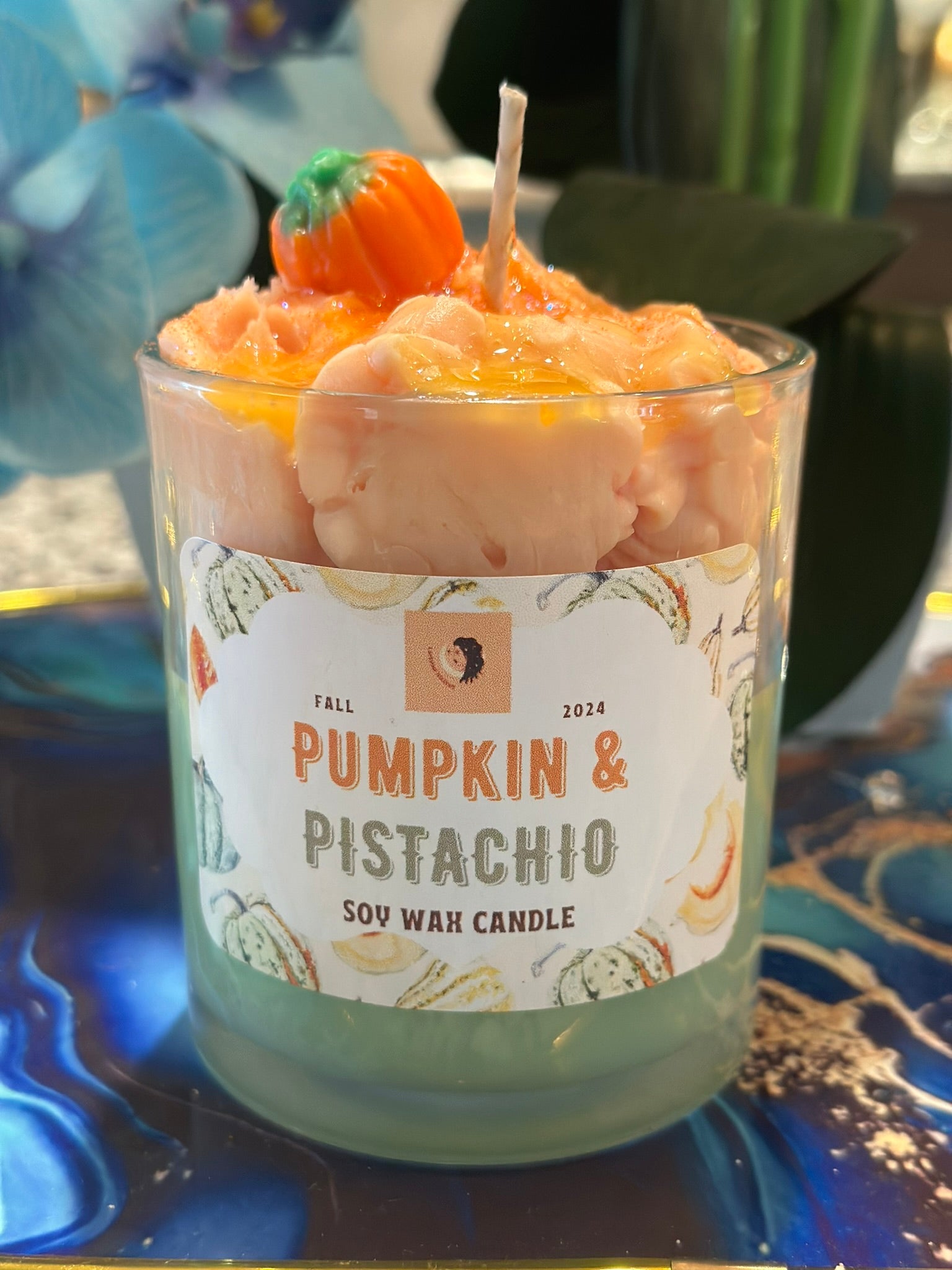 A glass jar candle labeled 'Pumpkin & Pistachio' with a white paper label, filled with pale pink wax and topped with a small orange pumpkin decoration, set on a blue marbled surface.