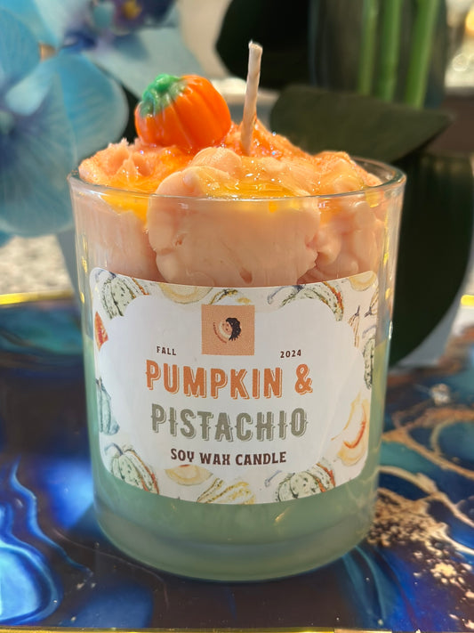 A glass jar candle labeled 'Pumpkin & Pistachio' with a white paper label, filled with pale pink wax and topped with a small orange pumpkin decoration, set on a blue marbled surface.