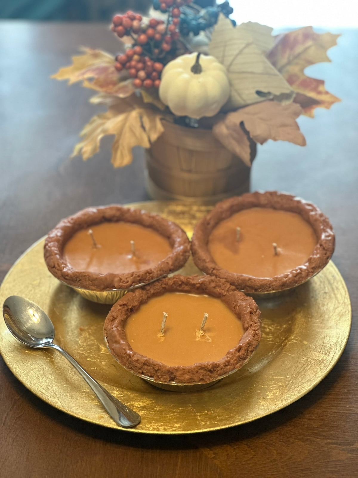 Three small sweet potato pie candles on a gold plate with a decorative arrangement in the background.