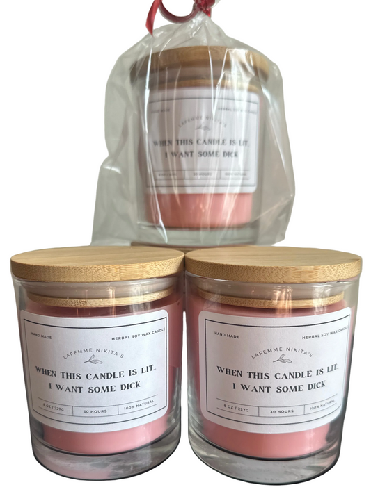 Three adult titled soy wax candles in clear glass jars with gold lids are shown on a white background, including two in front and one in a clear drawstring bag behind.