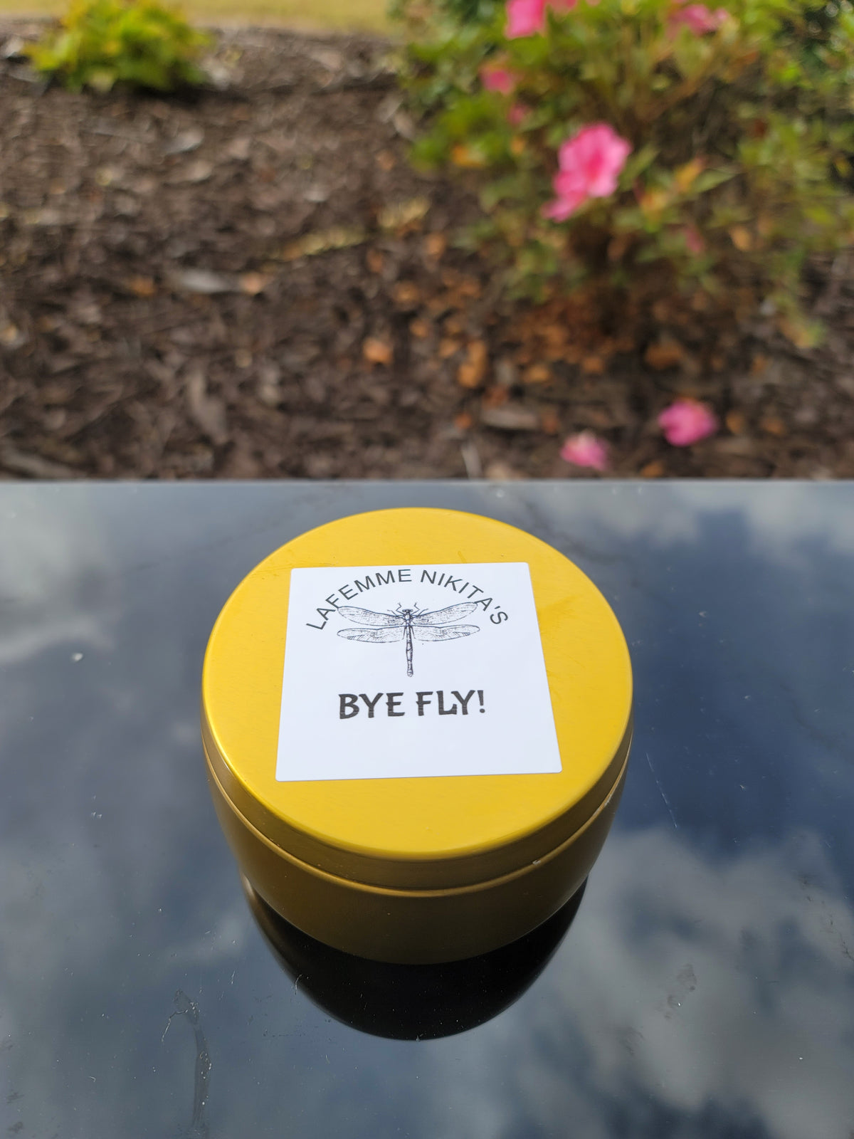 Gold container citronella candle with a sticker on a reflective surface with a garden background