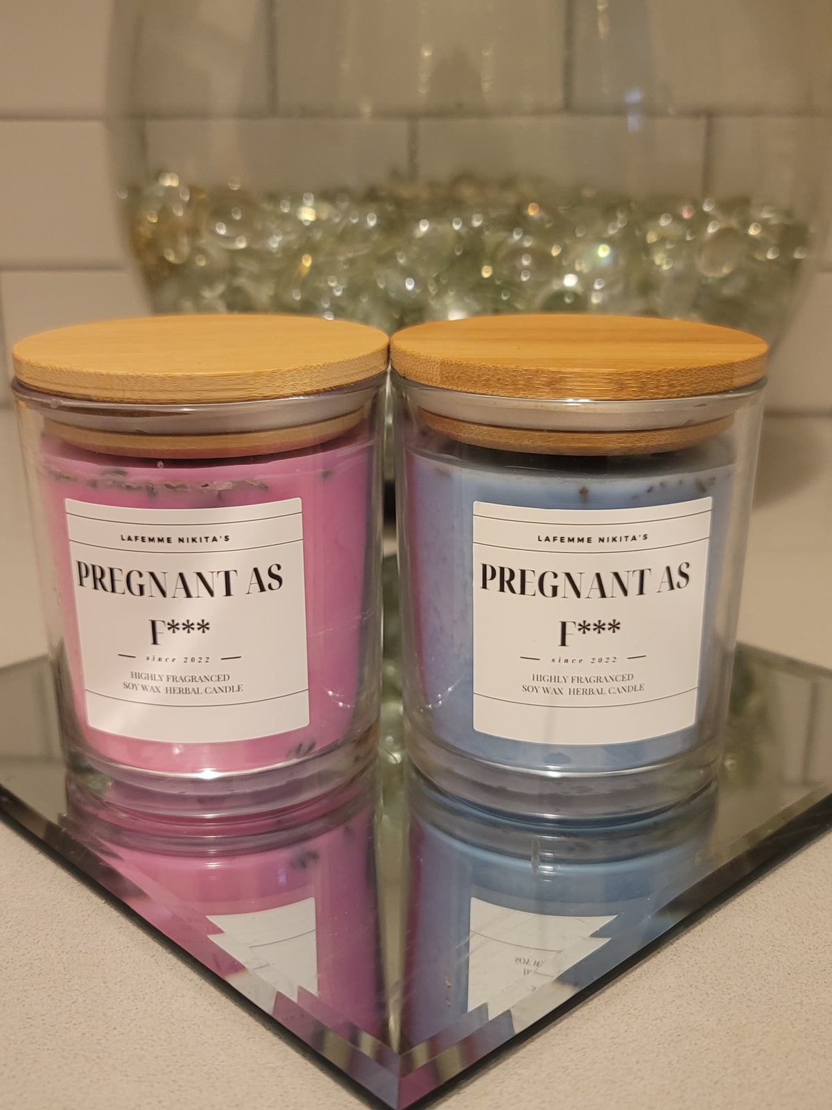 Two candles with clear glass jars, one labeled 'Pregnant As F***' with a pink lid and the other with a blue lid, reflecting in a mirror.